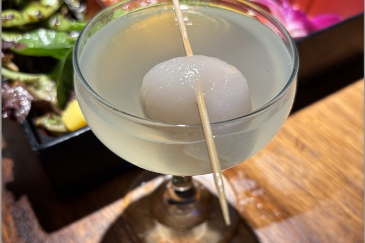 Cocktail with a lychee garnish on a skewer, bento box in the background.