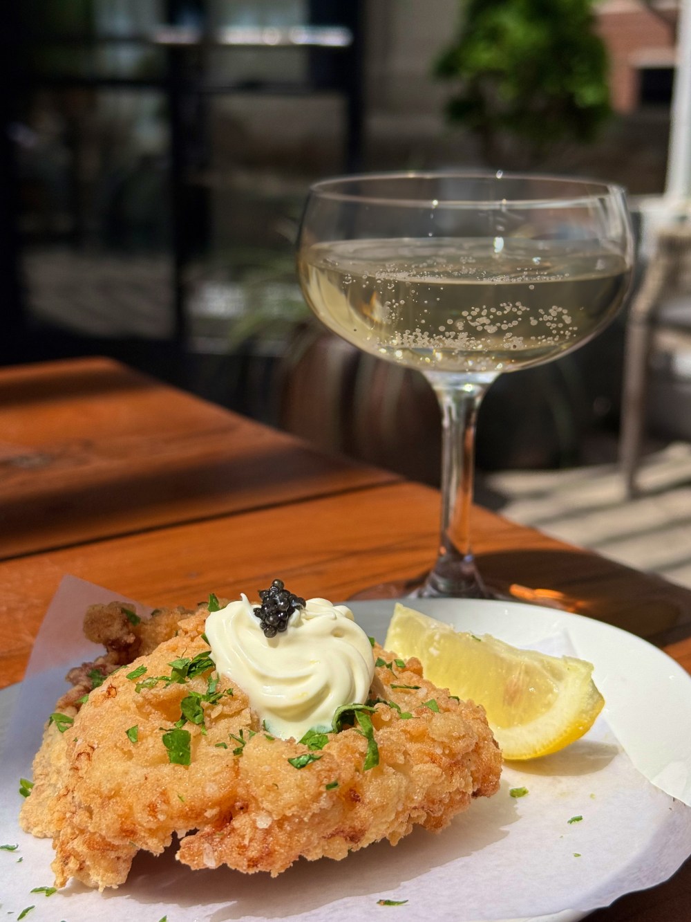Fried chicken with lemon wedge, topped with sauce; glass of sparkling wine on wooden table.